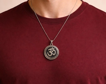 May include: Silver-toned pendant necklace featuring the Om symbol. The circular pendant has a dark background with the Om symbol in the center. The necklace is worn on a maroon t-shirt.