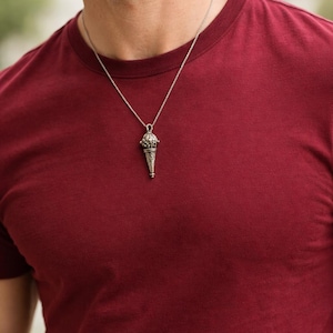 May include: A silver pendant necklace with a detailed, cone-shaped charm. The necklace is worn over a maroon t-shirt. The charm has intricate patterns and a decorative top. The background is blurred, focusing on the necklace and shirt.