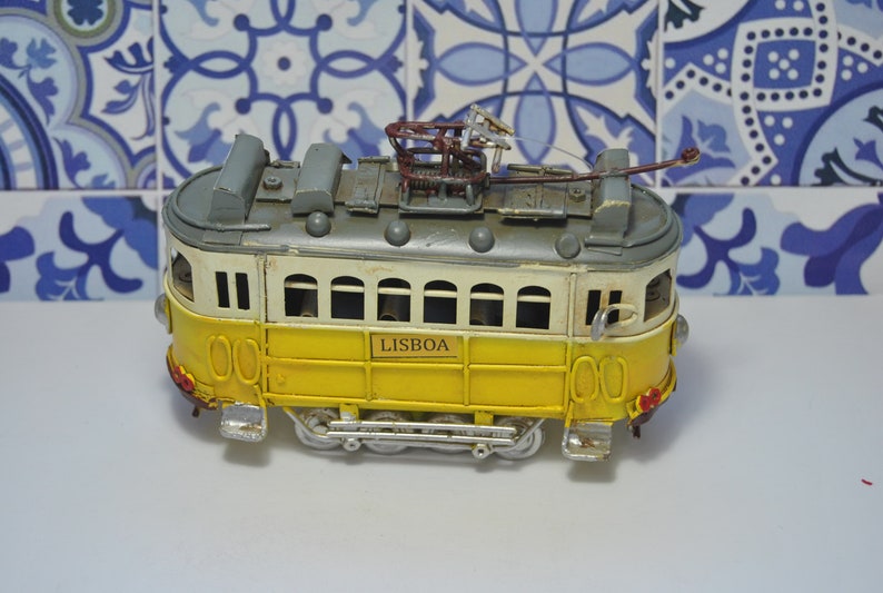 May include: A yellow and white toy model of a Lisbon tram with the word "LISBOA" printed on the side. The tram has a gray roof and a metal trolley pole.