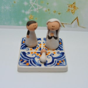 May include: A ceramic nativity scene with a white background and blue and orange accents. The figures are made of white clay and have painted details. The scene includes Mary, Joseph, and the baby Jesus.