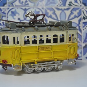 May include: A yellow and white toy model of a Portuguese tram with the word "PORTUGAL" printed on the side. The tram has a gray roof and a metal construction.