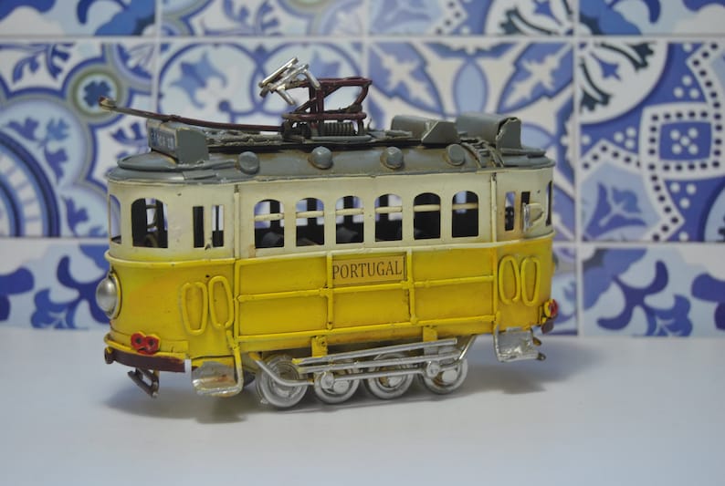 May include: A yellow and white toy model of a Portuguese tram with the word "PORTUGAL" printed on the side. The tram has a gray roof and a metal construction.