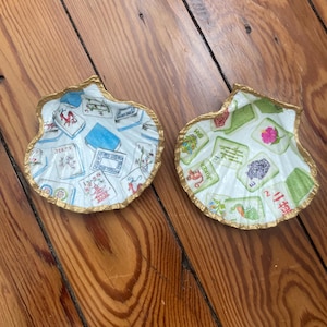 May include: Two decorative seashell-shaped dishes. The interior of each shell is decorated with a colorful pattern of Mahjong tiles. The edges are trimmed with gold paint. The shells are resting on a wooden surface.