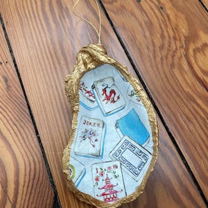 May include: A decorative oyster shell ornament with a gold-painted rim and a gold string for hanging. The interior features a collage of colorful Mahjong tiles. The ornament is set against a wooden background.