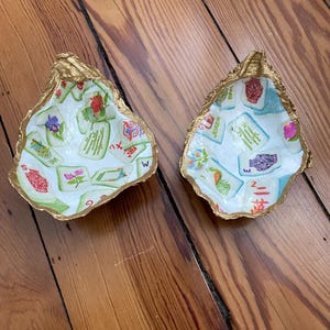 May include: Two decorative oyster shells with gold-painted edges. The interior of each shell features a white background decorated with a colorful collage of Mahjong tiles, including various floral patterns, Chinese characters, and a Joker tile.