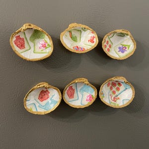 Puede incluir: Seis conchas marinas individuales con bordes pintados en dorado, cada una con un diseño interior de decoupage único inspirado en fichas de mahjong. Los patrones incluyen ilustraciones coloridas de jarrones, flores y la palabra Joker sobre fondo blanco.