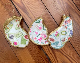 Op de afbeelding: Drie oesterschelp-ornamenten met goudkleurige randen en gouden ophanglusjes. De binnenzijden zijn voorzien van een decoupage-patroon van kleurrijke mahjongtegels, waaronder bloemmotieven, Chinese karakters en joker-tegels op een witte ondergrond.