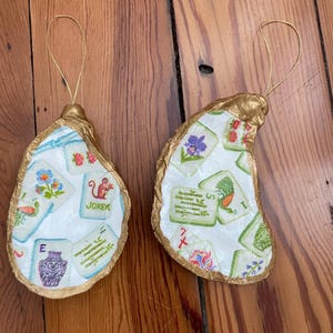 May include: Two oyster shell ornaments with gold-painted edges and gold hanging strings. The shells feature a decoupage pattern of Mahjong tiles, including a Joker tile with a monkey, floral designs, bamboo, and a purple vase on a white background.