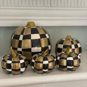May include: A collection of five decorative pumpkins featuring a hand-painted black, white, and metallic gold checkered pattern. Each pumpkin is topped with a stem covered in sparkling gold glitter, creating a cohesive set of varying sizes.