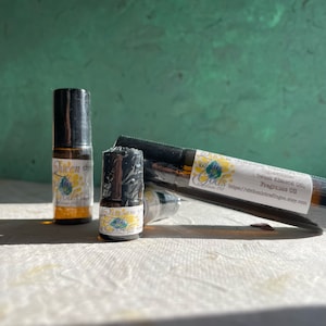 Puede incluir: Tres pequeños frascos de vidrio con tapas de rodillo negras. Los frascos contienen un líquido transparente y tienen etiquetas con el texto "Queen of Gods" y "Fragrance Oil".