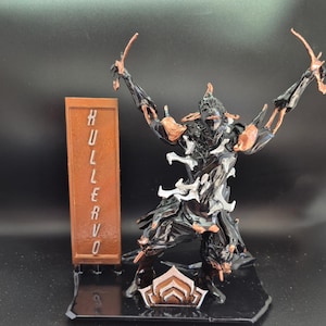 May include: A black and silver 3D printed figurine of a character from the video game Warframe. The figurine is posed with arms raised and is standing on a black base with a copper-colored symbol. The figurine is labeled "HULLER VO" on a copper-colored plaque.