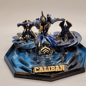 May include: A blue and gold 3D printed figurine of a character from the video game Warframe. The character is named "Caliban" and is standing on a blue and black base.