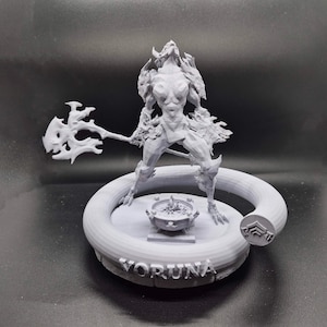 Può includere: Una statuetta stampata in 3D di una guerriera femminile che tiene un'ascia grande. La statuetta è in piedi su una base circolare con la scritta "VORUNA" incisa su di essa.