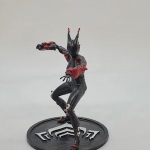 Può includere: Una statuetta nera e rossa di una figura umanoide stilizzata in posa dinamica. La figura è su una base circolare nera con un emblema bianco. La figura ha accenti rossi su braccia, gambe e testa.