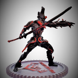 Può includere: Una action figure stampata in 3D di colore nero e rosso di un guerriero che tiene una spada. La figura è in piedi su una base grigia a forma di ingranaggio con la parola "WARFRAME" stampata su di essa.