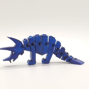 May include: A blue articulated dinosaur toy made of wood. The dinosaur is posed in a walking position with its head down. The toy is made of multiple pieces that are connected by hinges.