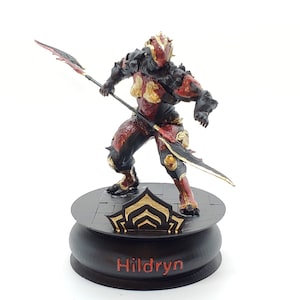 Può includere: Una statuetta stampata in 3D di un personaggio del videogioco Warframe. Il personaggio è Hildryn, una Warframe femminile con un design di armatura rosso e nero. Tiene una lancia e sta in piedi su una base nera con un simbolo dorato e il nome "Hildryn" scritto su di essa.