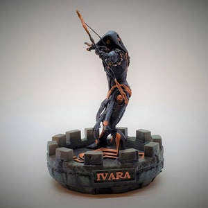Può includere: Una statuetta stampata in 3D di colore nero e oro che rappresenta una figura incappucciata che tiene un arco e una freccia. La statuetta è in piedi su una base verde e marrone con la scritta "IVARA" incisa su di essa.