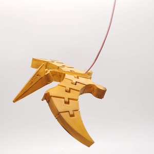 May include: A yellow wooden toy pterodactyl hanging from a pink string. The toy is made of interlocking pieces and has a long beak.