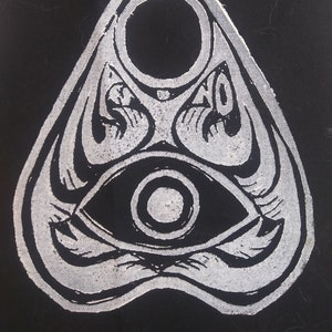 May include: A black and white planchette featuring a central eye graphic, a circular cutout at the top, and the words YES and NO printed on either side of the design. The object has a pointed top and a curved base with intricate line work.
