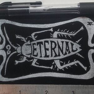 Puede incluir: Parche bordado en blanco y negro con un diseño de cucaracha y la palabra "ETERNAL" en una fuente estilizada. El parche mide aproximadamente 5 cm de ancho.