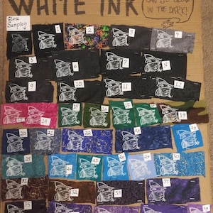 May include: A collection of fabric swatches with a white ink design of a frog wearing a hat. The swatches are numbered 1 through 39 and are arranged in rows on a cardboard background. The text "WHITE INK" is written at the top of the board. The text "CAN BE GLOW IN THE DARK!" is written in a speech bubble above the swatches. The text "Glow Samples" is written above the first row of swatches.