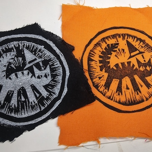 May include: Two fabric patches with a black and white circular design. The design is a stylized creature with a large mouth and sharp teeth. The patches are on a white background.