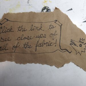 May include: A brown paper scroll with the text "Click the link to see close-ups of all of the fabric!" written in black ink. A cartoon drawing of a crowned wolf's head is on the right side of the scroll.