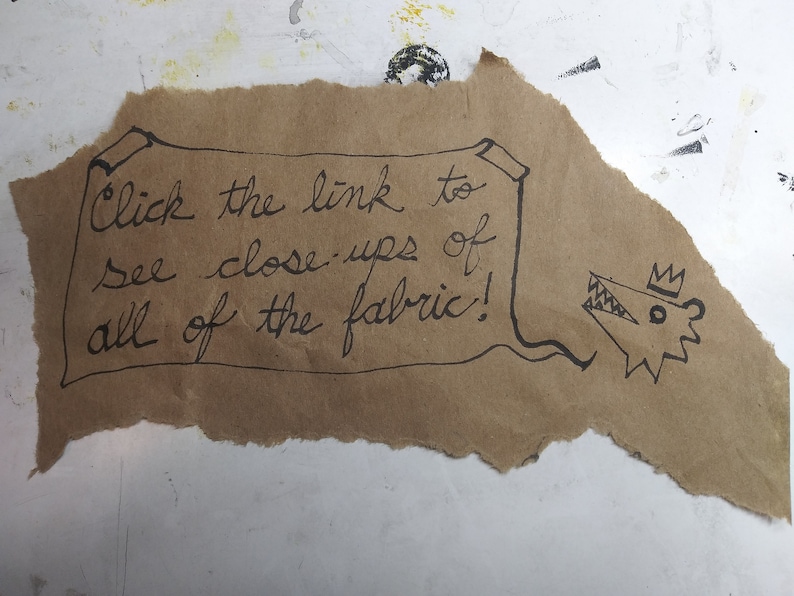 May include: A brown paper scroll with the text "Click the link to see close-ups of all of the fabric!" written in black ink. A cartoon drawing of a crowned wolf's head is on the right side of the scroll.