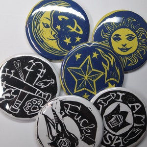 May include: Six round, black and white, and blue and yellow buttons with various designs. The designs include a crescent moon, a sun, a star, a skull, a bird, and a symbol with a cross and a paw print.