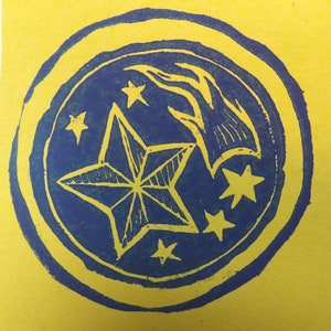 Puede incluir: Una impresión azul y amarilla de una estrella de cinco puntas con un diseño de llama estilizado detrás. Hay cinco estrellas más pequeñas que rodean la estrella grande.