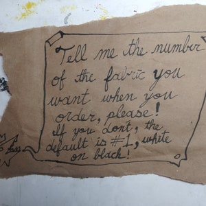 May include: A handwritten note on brown paper with the text: "Tell me the number of the fabric you want when you order, please! If you don't, the default is #1, white on black!"
