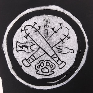May include: A white circle with a black background, featuring a graphic of crossed baseball bats, a knuckle duster, and other weapons. The graphic is in a white outline style.