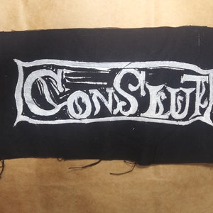 Puede incluir: Un parche rectangular negro con la palabra "CONSTITUT" impresa en blanco con letras estilizadas. El parche tiene un borde deshilachado y probablemente está hecho de tela. El fondo es de color marrón claro.