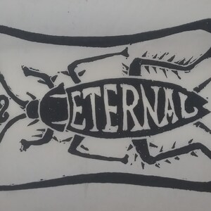 May include: Black and white linocut print featuring a stylized insect with the word "ETERNAL" in the center. The insect is surrounded by a decorative border with swirling elements. The artwork is on a white background.