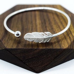 The bracelet is open-ended with a small silver ball at one end and a detailed feather charm at the other. The feather charm is intricately designed, with fine lines representing the texture of a real feather.