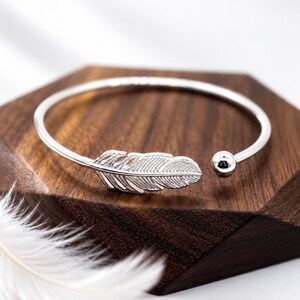 Könnte beinhalten: Ein silberfarbenes, offenes Armband mit einem detaillierten Federanhänger an einem Ende und einer glatten, polierten kugelförmigen Perle am anderen Ende. Das Metallband ist dünn und kreisförmig und weist ein reflektierendes Finish auf.