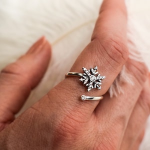 Puede incluir: Un anillo de plata con un diseño de copo de nieve con pequeños diamantes de imitación transparentes. El anillo es abierto y se envuelve alrededor del dedo.