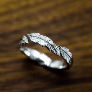 May include: A silver ring with a feather design. The ring has a detailed feather pattern that goes around the band. The ring is displayed on a wooden surface. The ring is a piece of jewellery.