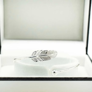 May include: A silver-toned open cuff bracelet featuring a detailed feather design on one end and a small spherical bead on the other. The bracelet is displayed inside a white jewelry box with a black frame, resting on a white velvet insert.