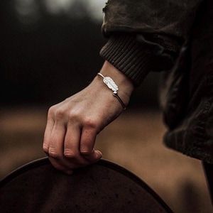 May include: A silver-toned metal cuff bracelet featuring a delicate feather charm and a small spherical bead at the end. The bracelet is worn on the wrist, showcasing a polished finish and a slender, open-ended design that curves around the arm.