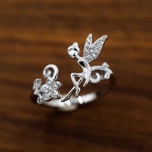 May include: Silver ring featuring a fairy perched on a branch with a flower detail. The fairy and flower are adorned with small clear stones.