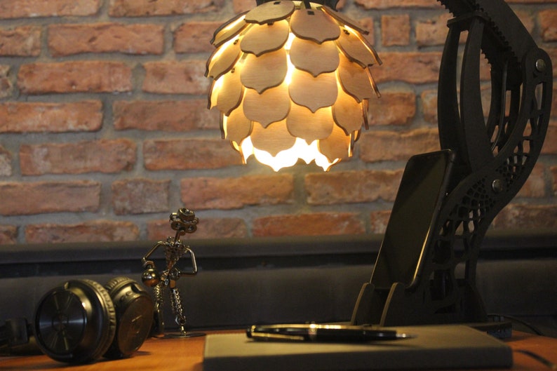 May include: A black desk lamp with a unique, articulated arm and a wooden, geometric shade. The lamp is on a desk with a phone, headphones, and a pen.
