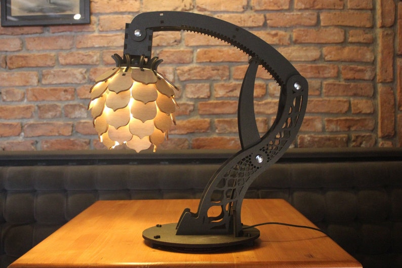 May include: A black, articulated lamp with a wooden, geometric shade. The lamp is on a wooden table and has a black cord.