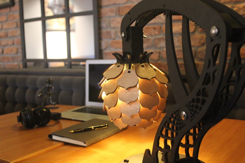 May include: A black metal lamp with a geometric design and a wooden pineapple-shaped shade. The lamp is on a wooden table with a laptop, a notebook, and a pen.