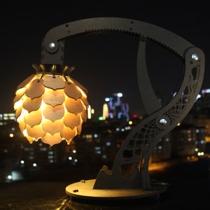 May include: A wooden lamp with a unique design resembling a robotic arm holding a light fixture shaped like a pine cone. The lamp is illuminated and casts a warm glow. The background is a blurry cityscape at night.