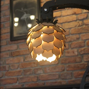 May include: A wooden lampshade shaped like a pine cone with a warm light shining through the cutouts. The lampshade is attached to a black metal arm.