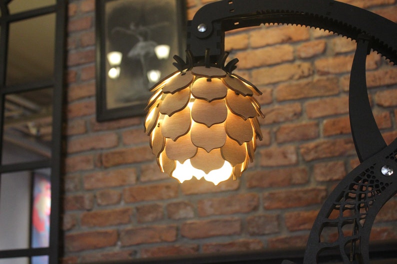 May include: A wooden lampshade shaped like a pine cone with a warm light shining through the cutouts. The lampshade is attached to a black metal arm.