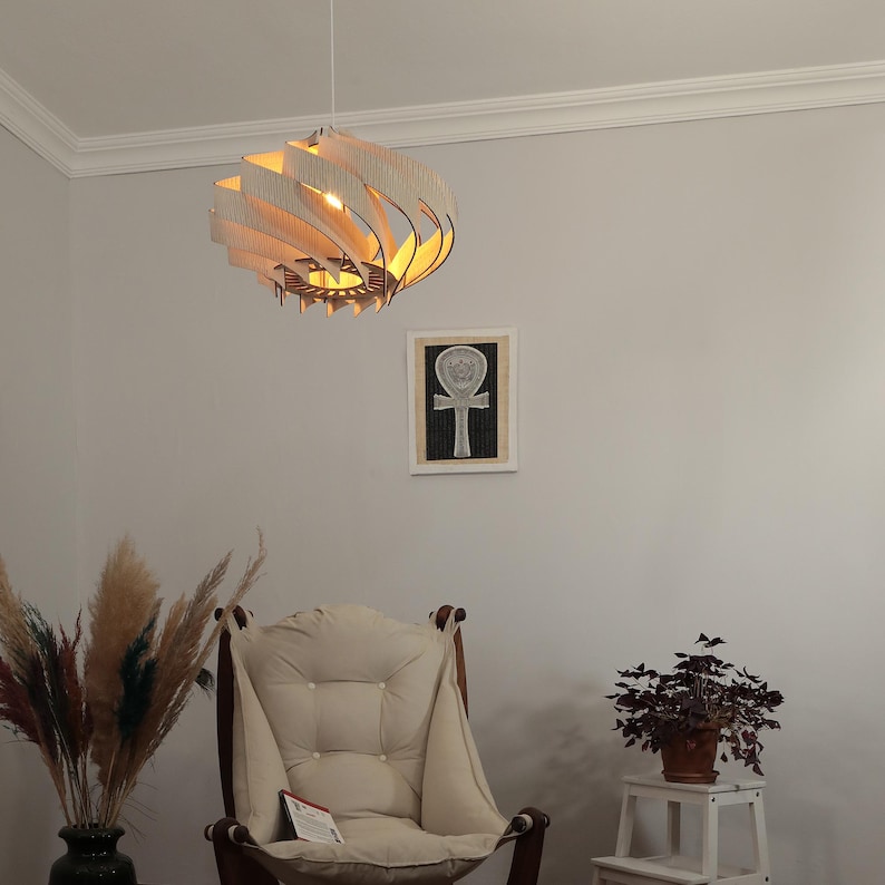 May include: A white wooden pendant light with a unique, swirling design hangs from the ceiling. The light is on and casts a warm glow. A white armchair with a thick cushion sits in front of a white wall. A small potted plant sits on a white stool.