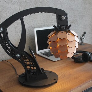 May include: A black desk lamp with a unique, articulated arm and a wooden, geometric shade that resembles a pine cone. The lamp is on a wooden desk with a laptop, headphones, and a notebook.